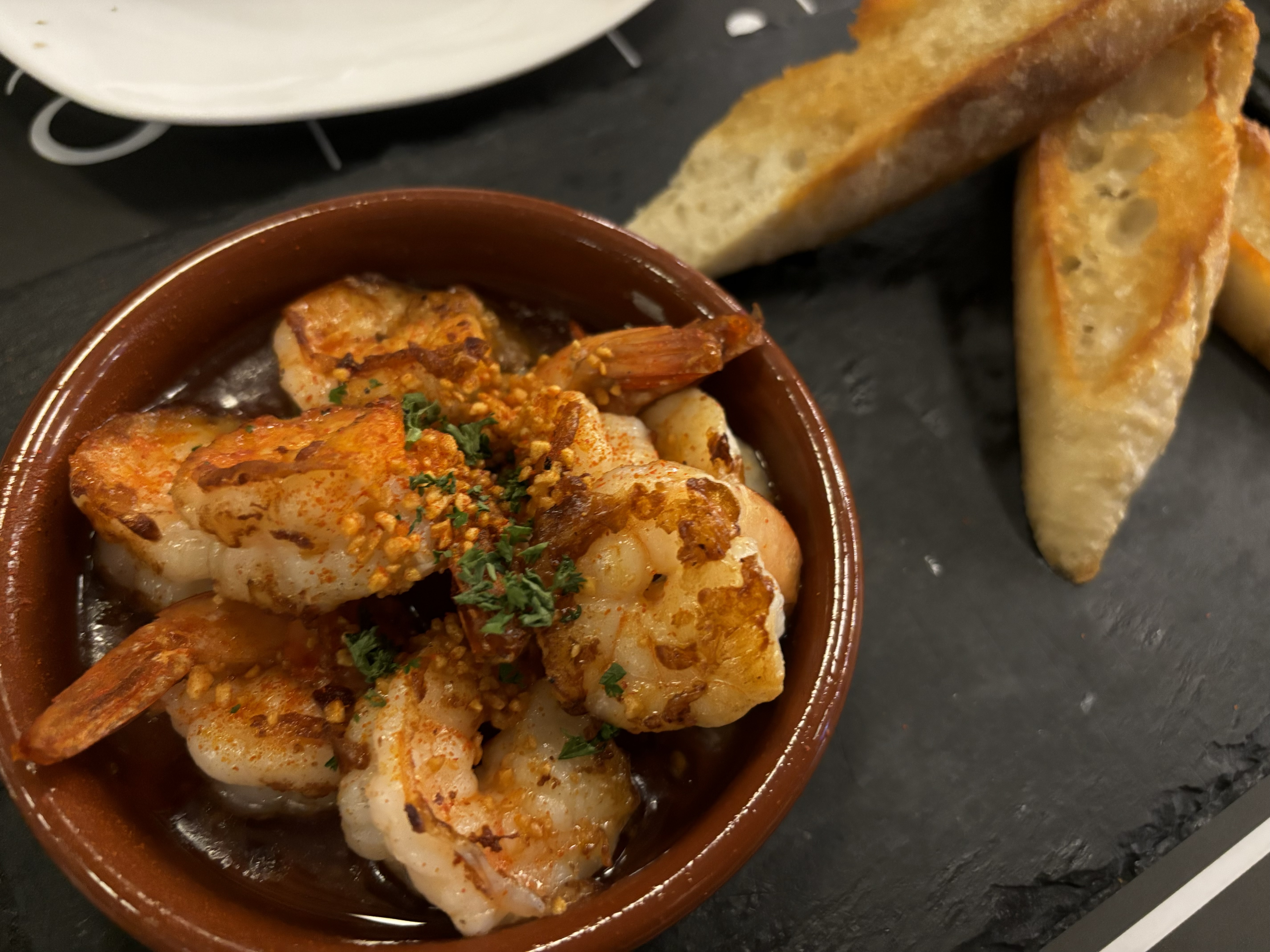 Shrimp in olive oil with garlic and chili in a small pan, parsley, bread beside, Natural window light, ivory linen, handmade ceramic, shallow depth of field, no text, no logo, no watermark, fine-dining magazine photograph. Spanish Garlic Shrimp